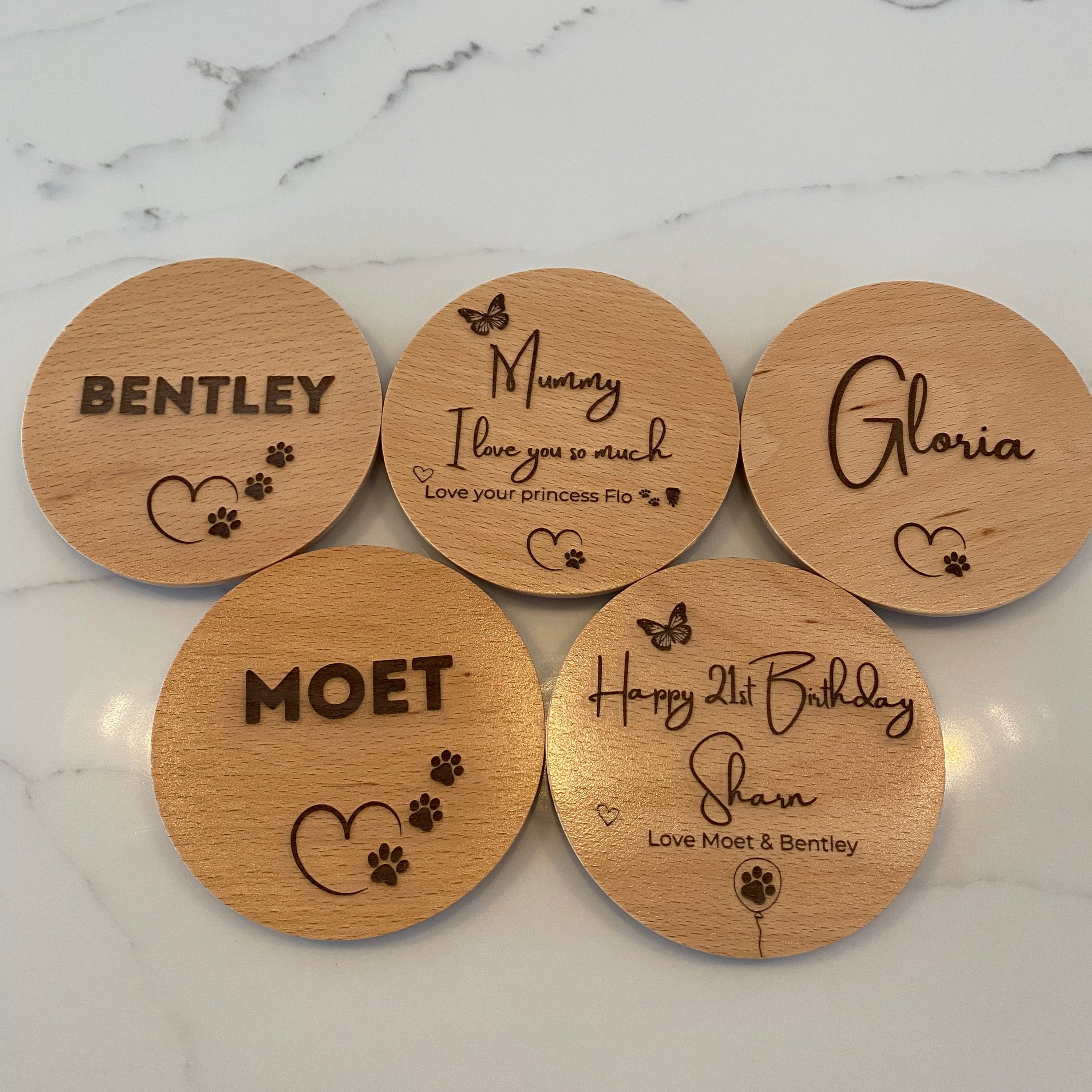 Five personalised wooden candle lids with engraved names and paw print designs, arranged on a white marble surface.