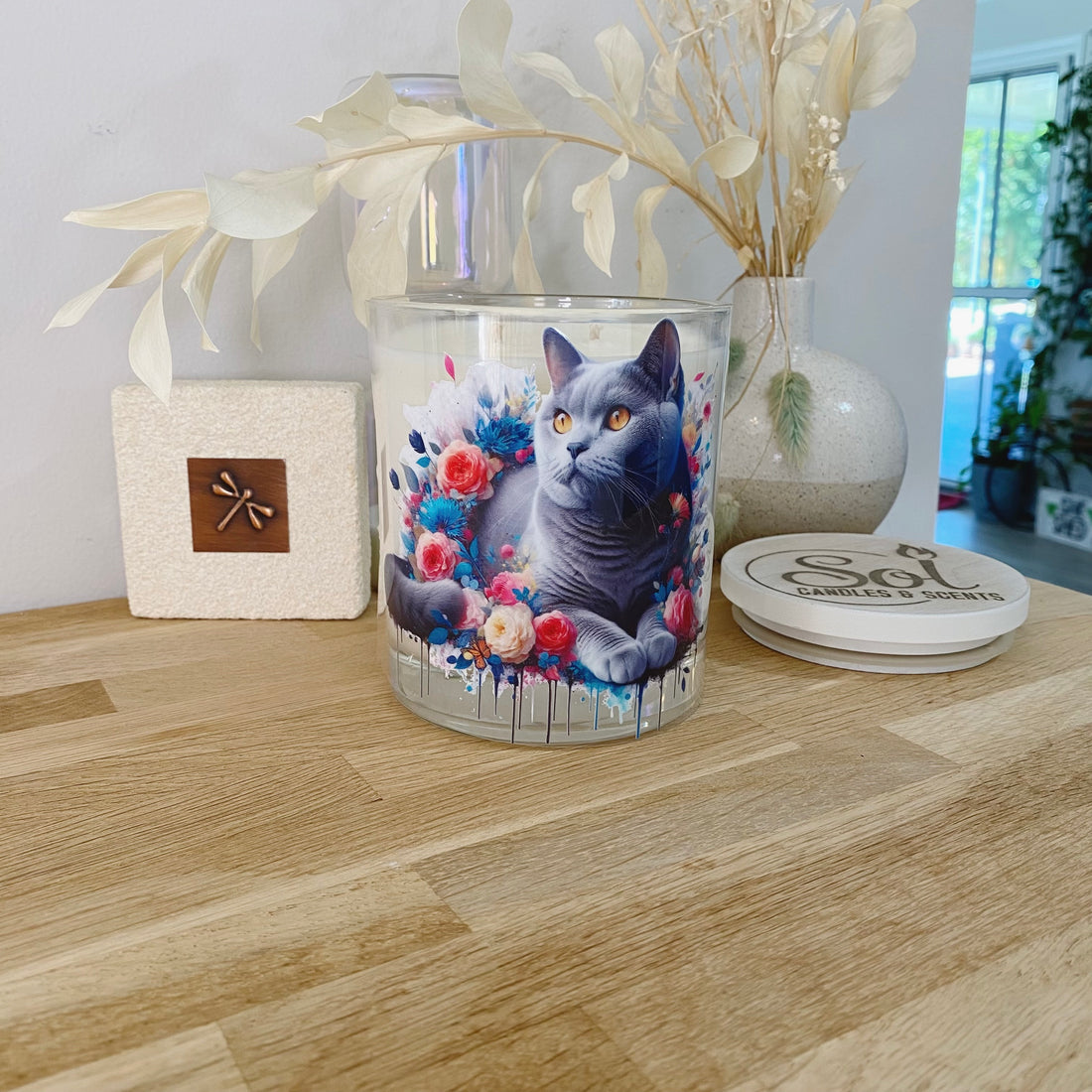 Glass candle with a cat design on a wooden surface, surrounded by decorative items.