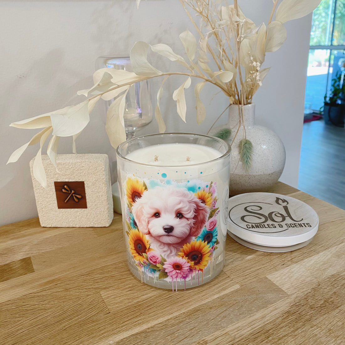 Candle with a Cavoodle dog design on a wooden surface next to a vase with dried leaves and a coaster.