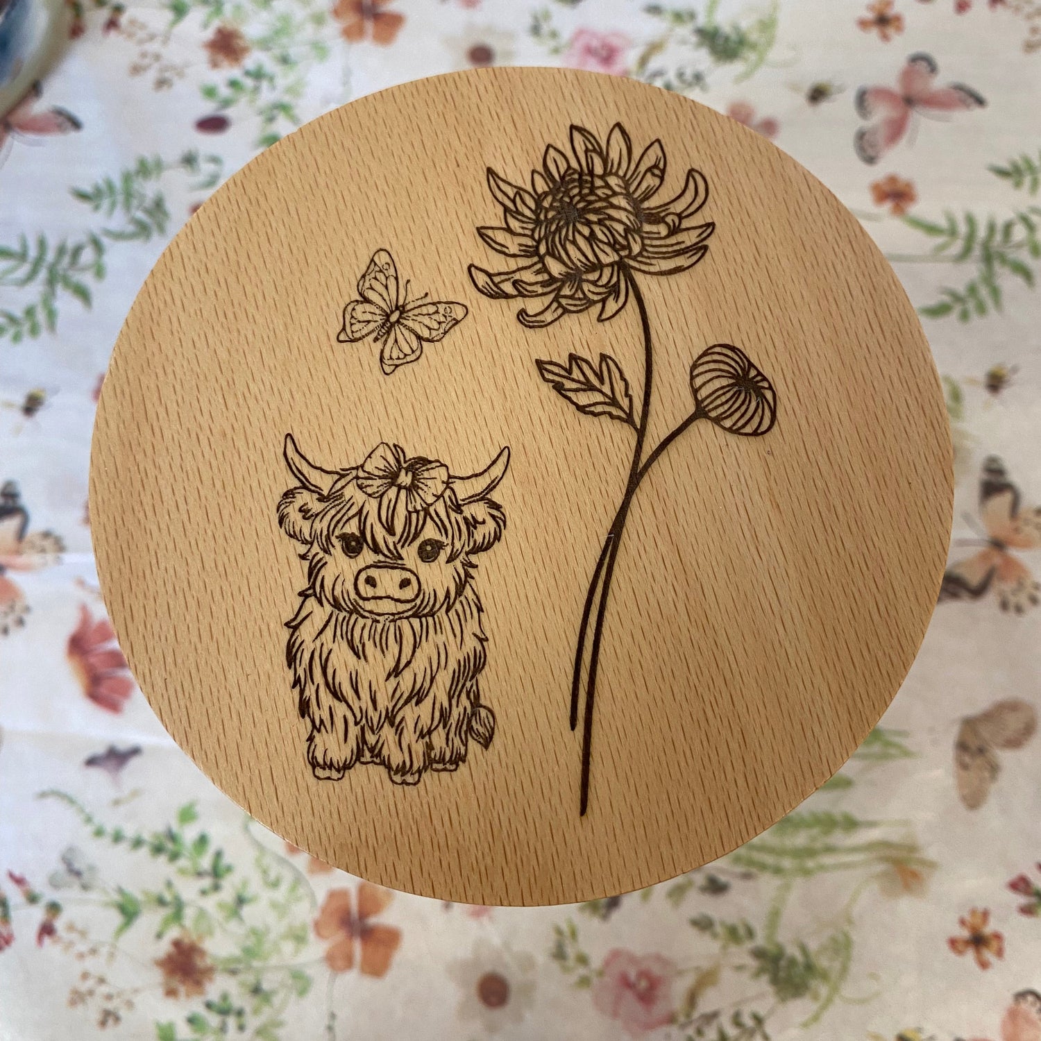 Highland Mini Cow illustration with a flower and butterfly on a wooden surface, highlighting the Highland Mini Cow design.