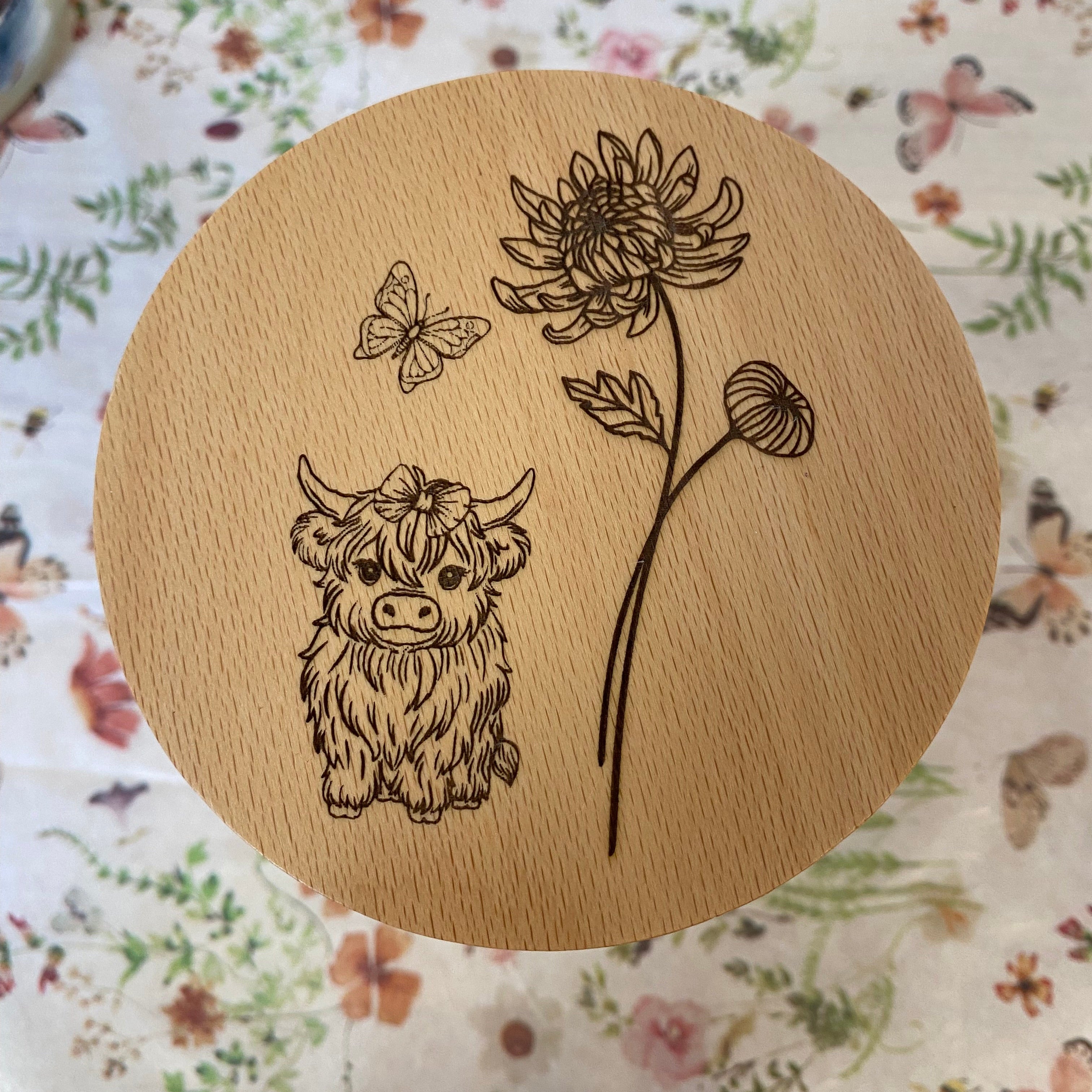 Highland Mini Cow illustration with a flower and butterfly on a wooden surface, highlighting the Highland Mini Cow design.