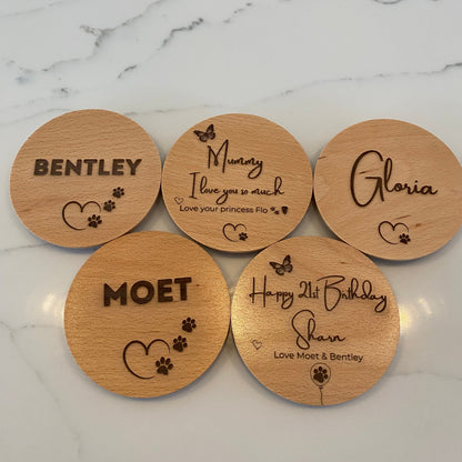 Five personalised wooden candle lids with engraved names and paw print designs, arranged on a white marble surface.